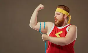 Rudowłosy mężczyzna typu endomorfik w żółtej opasce i czerwonej koszulce mierzy obwód bicepsa.