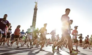 Biegacze w maratonie, z Kolumną Zwycięstwa w tle. Słońce świeci, tworząc efekt "negative split" na twarzach niektórych zawodników.