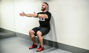 Mężczyzna z brodą wykonuje ćwiczenie wall sit, opierając plecy o ścianę i uginając nogi w kolanach.