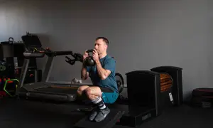 Kolarz wykonuje głęboki przysiad z kettlebell. W tle bieżnia i sprzęt do ćwiczeń.