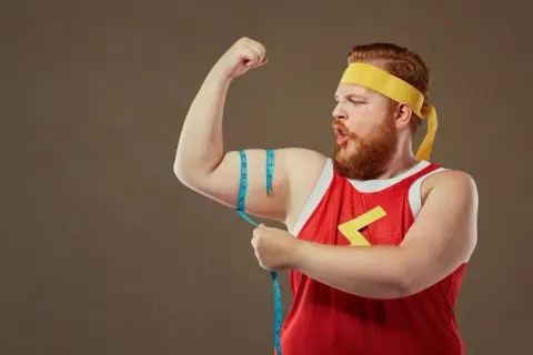Rudowłosy mężczyzna typu endomorfik w żółtej opasce i czerwonej koszulce mierzy obwód bicepsa.