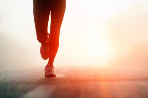 Biegaczka w świetle wschodzącego słońca. Jej nogi w legginsach i butach sportowych są w ruchu. To jak test coopera, gdzie każdy krok się liczy.