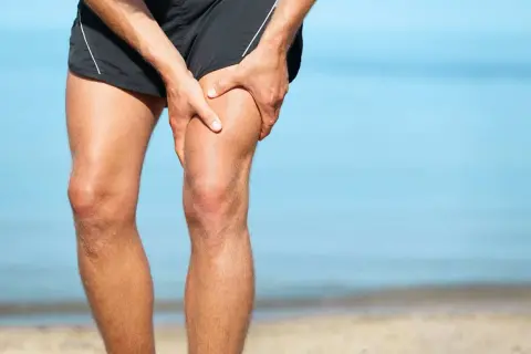 Ból w udzie, gdzie znajduje się mięsień czworogłowy. Mężczyzna trzyma się za nogę na plaży.