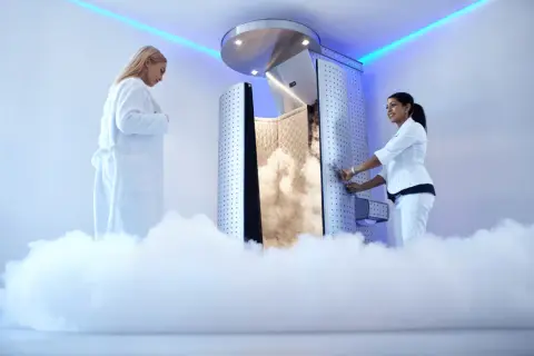 Kobieta w białym szlafroku czeka na zabieg krioterapii. Obok niej pracownica otwiera drzwi komory wypełnionej mgłą.