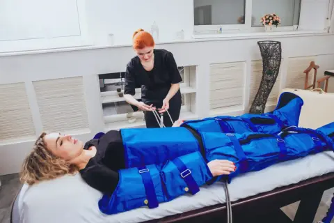 Kobieta w salonie piękności leży na łóżku, ubrana w niebieski kombinezon do drenażu limfatycznego. Terapeutaka przygotowuje zabieg, by zapewnić widoczne efekty.