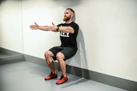 Mężczyzna z brodą wykonuje ćwiczenie wall sit, opierając plecy o ścianę i uginając nogi w kolanach.