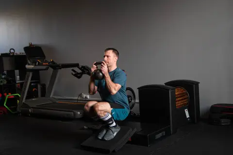 Kolarz wykonuje głęboki przysiad z kettlebell. W tle bieżnia i sprzęt do ćwiczeń.