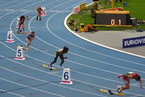 Biegaczki na 400 m walczą o rekord świata. Na bieżni widać zawodniczki w akcji, numery torów i zegar z wynikiem 0.1.
