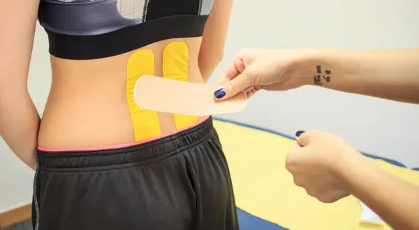 Naklejanie tejpy na plecy w odcinku lędźwiowym. Dłoń z tatuażem "You can do it" przygotowuje plaster.