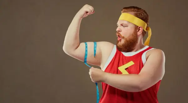 Rudowłosy mężczyzna typu endomorfik w żółtej opasce i czerwonej koszulce mierzy obwód bicepsa.