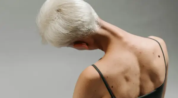 Widok z tyłu na osobę z krótkimi blond włosami, pochyloną głową. Widoczny mięsień dźwigacz łopatki i pieprzyki na plecach.