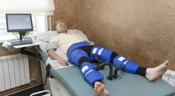Mężczyzna leży na łóżku rehabilitacyjnym, poddając się zabiegowi limfodrenaż. Na nogach ma założone specjalne mankiety.