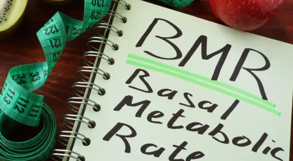 Notatnik z napisem BMR (Basal Metabolic Rate), obok jabłko, kiwi i miarka krawiecka. Obliczenia BMR, jak wzór Harrisa Benedicta, pomagają zrozumieć metabolizm.
