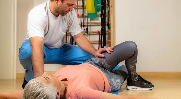 Fizjoterapeuta pomaga pacjentce wykonać test Patricka. Kobieta leży na macie, zgięte nogi w kolanach, a mężczyzna delikatnie naciska na jej kolano.