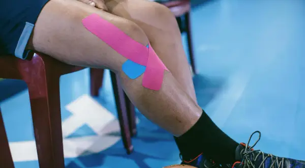 Osoba z chondromalacją rzepki stosuje kinesiotaping. Różowe i niebieskie plastry na kolanie wspierają ćwiczenia.