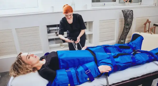 Kobieta w salonie piękności leży na łóżku, ubrana w niebieski kombinezon do drenażu limfatycznego. Terapeutaka przygotowuje zabieg, by zapewnić widoczne efekty.