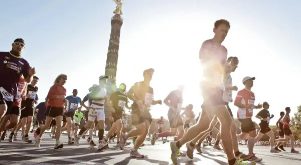 Biegacze w maratonie, z Kolumną Zwycięstwa w tle. Słońce świeci, tworząc efekt "negative split" na twarzach niektórych zawodników.