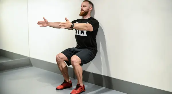 Mężczyzna z brodą wykonuje ćwiczenie wall sit, opierając plecy o ścianę i uginając nogi w kolanach.