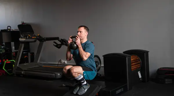 Kolarz wykonuje głęboki przysiad z kettlebell. W tle bieżnia i sprzęt do ćwiczeń.