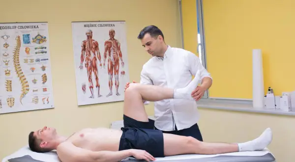 Fizjoterapeuta bada pacjenta z podejrzeniem naderwanego ścięgna. Na ścianie wiszą anatomiczne plakaty.