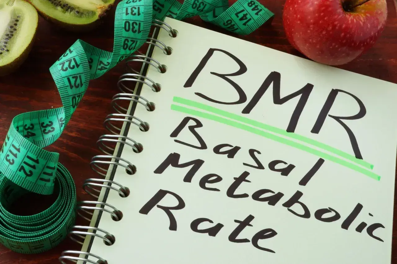 Notatnik z napisem BMR (Basal Metabolic Rate), obok jabłko, kiwi i miarka krawiecka. Obliczenia BMR, jak wzór Harrisa Benedicta, pomagają zrozumieć metabolizm.
