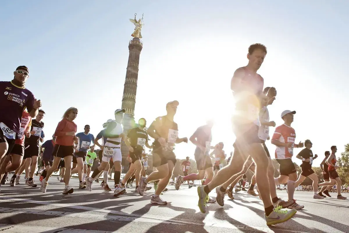 Biegacze w maratonie, z Kolumną Zwycięstwa w tle. Słońce świeci, tworząc efekt "negative split" na twarzach niektórych zawodników.