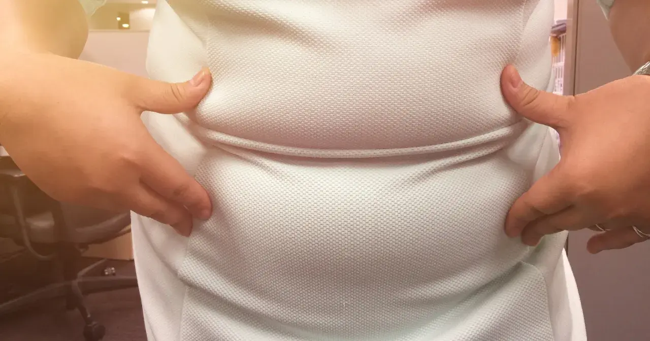Dłonie ściskają białą koszulkę, uwydatniając zaokrąglony brzuch, który może być oznaką brzucha hormonalnego.