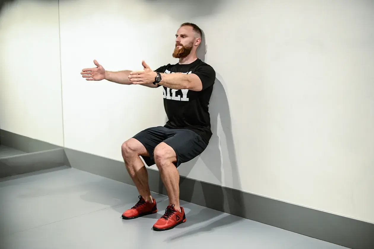 Mężczyzna z brodą wykonuje ćwiczenie wall sit, opierając plecy o ścianę i uginając nogi w kolanach.