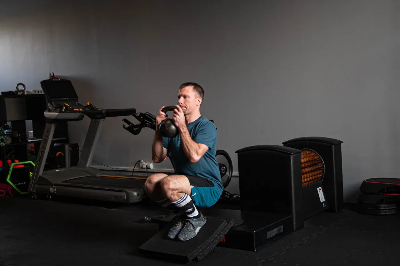 Kolarz wykonuje głęboki przysiad z kettlebell. W tle bieżnia i sprzęt do ćwiczeń.