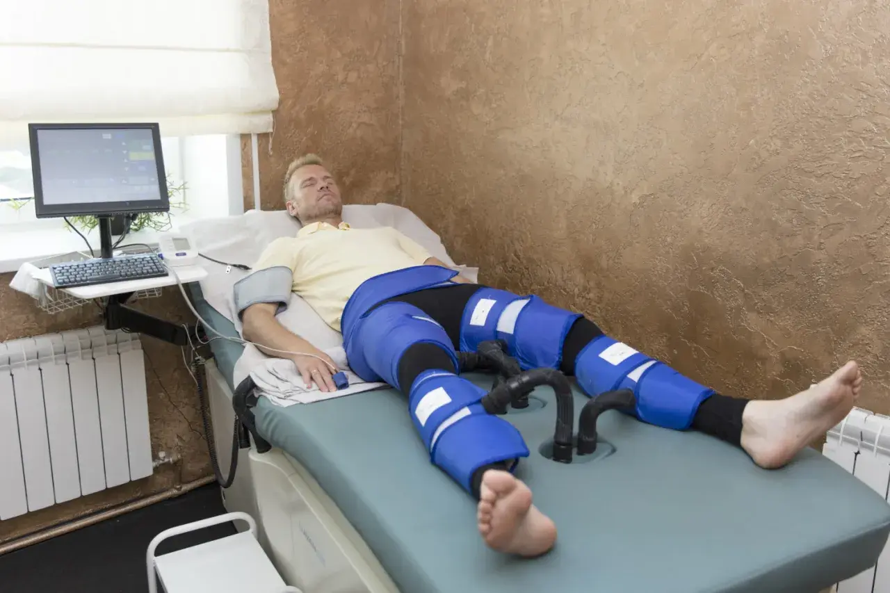 Mężczyzna leży na łóżku terapeutycznym, jego nogi są owinięte w niebieskie mankiety do drenażu limfatycznego. Monitor i sprzęt medyczny obok.