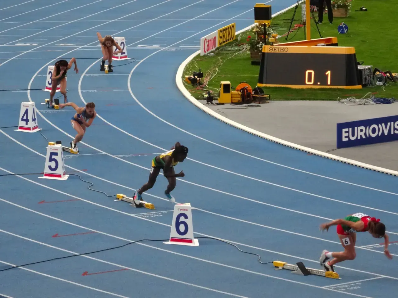 Biegaczki na 400 m walczą o rekord świata. Na bieżni widać zawodniczki w akcji, numery torów i zegar z wynikiem 0.1.
