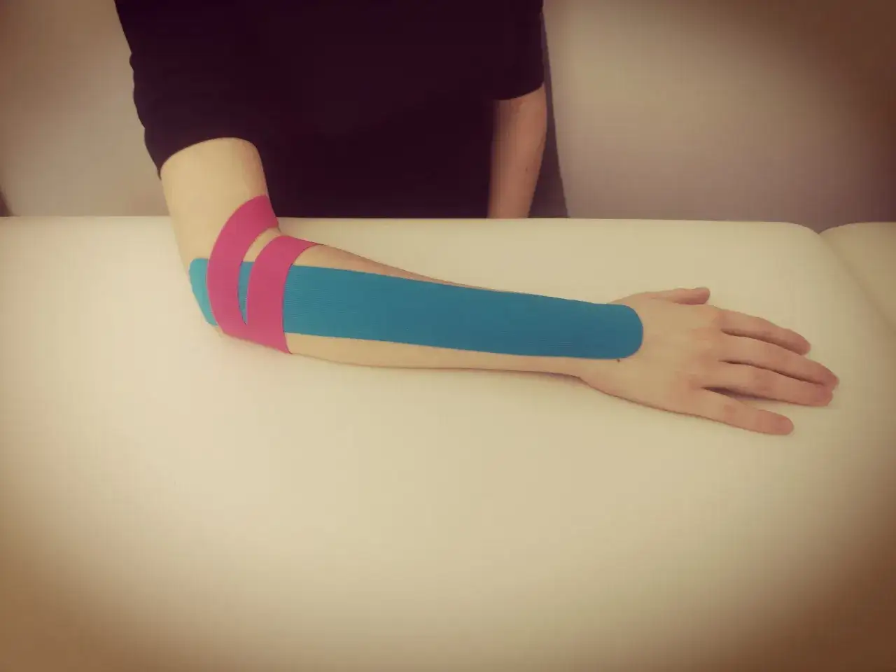 Ramię z aplikacją kinesiotaping na łokieć tenisisty. Taśmy w kolorze niebieskim i różowym wspierają mięśnie przedramienia, pomagając w rehabilitacji i ćwiczeniach.