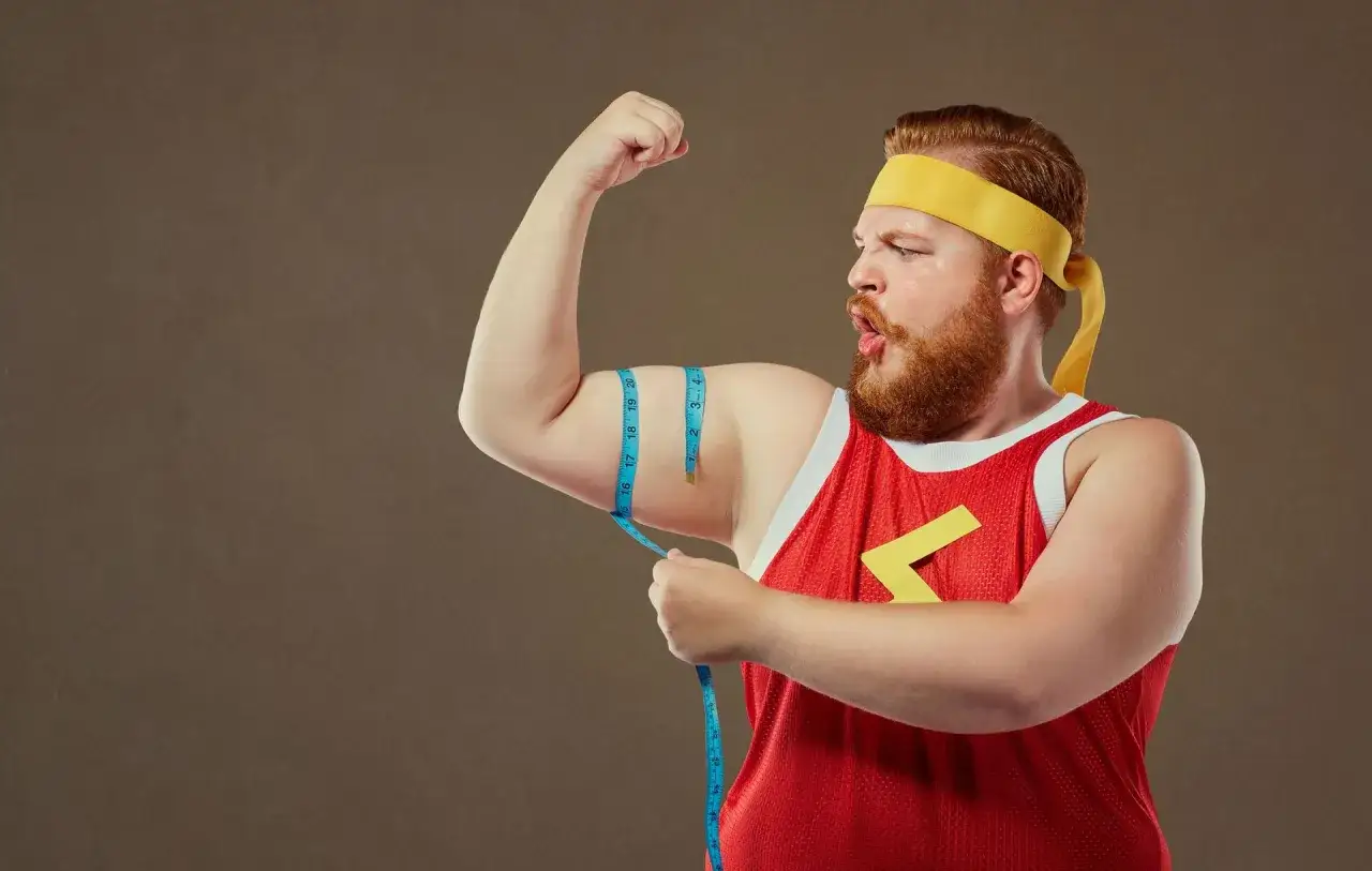 Mężczyzna typu endomorfik z wąsami i brodą, w żółtej opasce i czerwonej koszulce, mierzy obwód bicepsa.