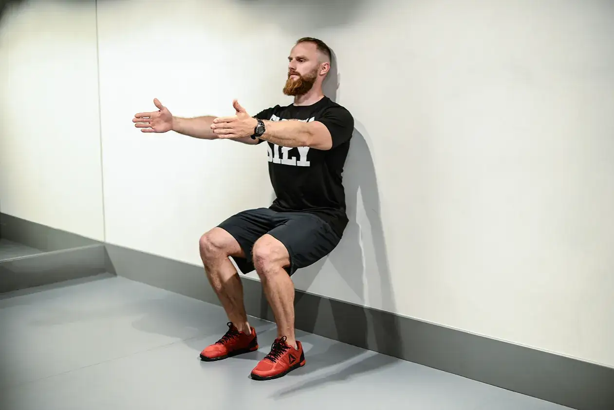Mężczyzna z brodą wykonuje ćwiczenie wall sit, opierając plecy o ścianę i uginając nogi w kolanach.