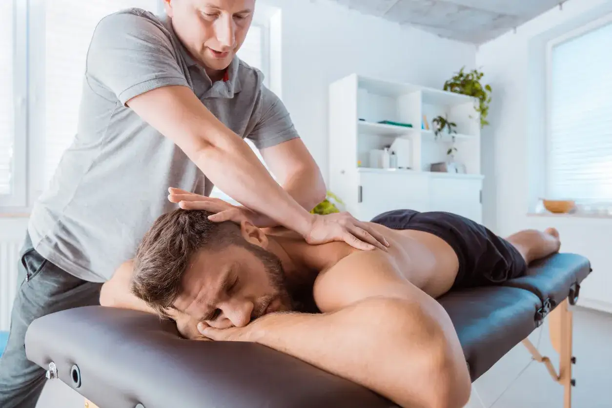 Mężczyzna leży na kozetce, a fizjoterapeuta wykonuje mu masaż sportowy pleców i karku.