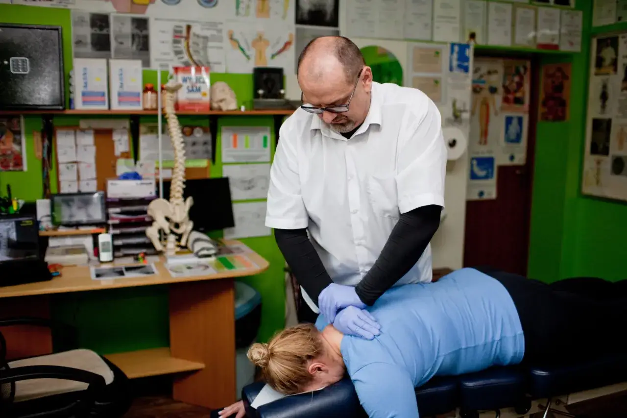 Chiropraktyka co to? Terapeuta wykonuje zabieg na karku pacjentki leżącej na kozetce. W tle model kręgosłupa i plakaty.