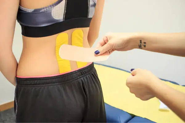 Naklejanie tejpy na plecy w odcinku lędźwiowym. Dłoń z tatuażem "You can do it" przygotowuje plaster.