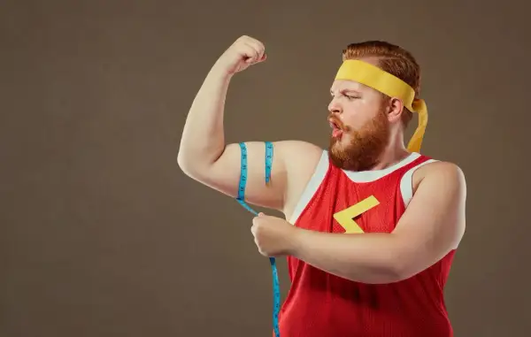 Rudowłosy mężczyzna typu endomorfik w żółtej opasce i czerwonej koszulce mierzy obwód bicepsa.