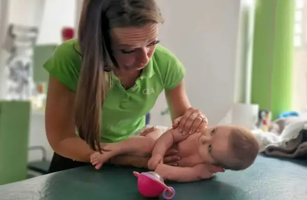 Fizjoterapeutka pracuje z niemowlęciem, pomagając mu w walce z **hipertonią**. Dziecko leży na zielonym stole, a kobieta delikatnie dotyka jego rączek.