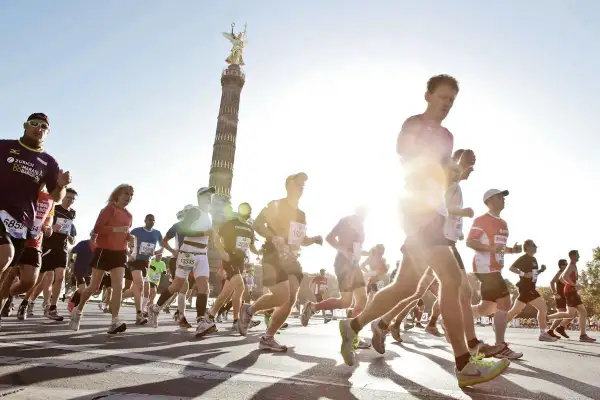 Biegacze w maratonie, z Kolumną Zwycięstwa w tle. Słońce świeci, tworząc efekt "negative split" na twarzach niektórych zawodników.